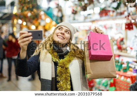 Smiling young woman spending time at Christmas fair and taking selfie on mobile phone