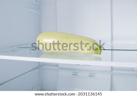 against the background of a white refrigerator, there is a zucchini on a glass shelf, close-up