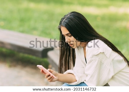 A girl writes SMS messages or uses applications on a smartphone while sitting on a bench in the park