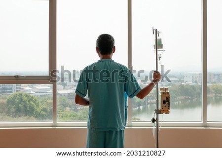 adult patient hold infusion hanger pole look out to hospital window, thinking about medical expenses and health care insurance alone, financial and wellness problem concept with copy space.