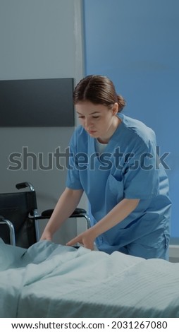 Caucasian nurse making hospital ward bed for use by sick person at modern medical facility unit. Emergency room with equipment, oxygen tube and wheelchair for intensive care treatment