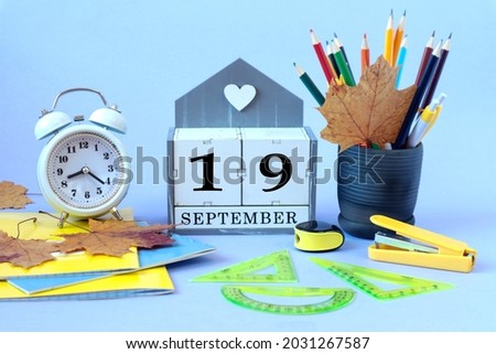 Calendar for September 19 : the name of the month of September in English, cubes with the number 19, clocks, school supplies, pencils and pens in a cup, maple leaves, blue background, side view