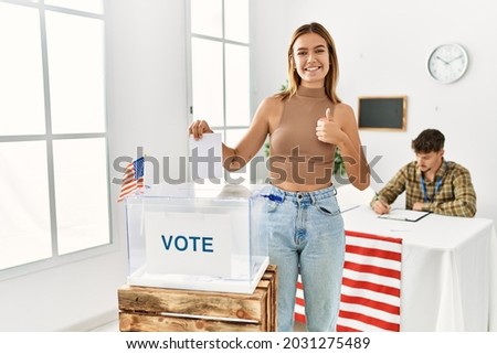 Young blonde girl voting putting envelop in ballot box smiling happy and positive, thumb up doing excellent and approval sign 