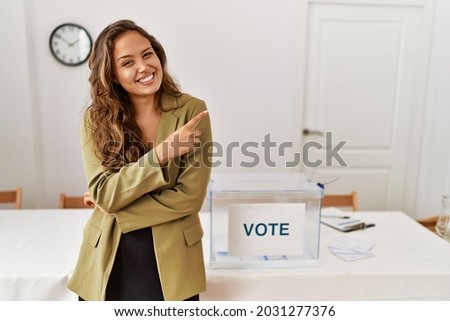 Beautiful hispanic woman standing at political campaign room with a big smile on face, pointing with hand and finger to the side looking at the camera. 