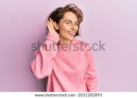 Young brunette woman wearing casual winter sweater smiling with hand over ear listening and hearing to rumor or gossip. deafness concept.  Royalty-Free Stock Photo #2031282893