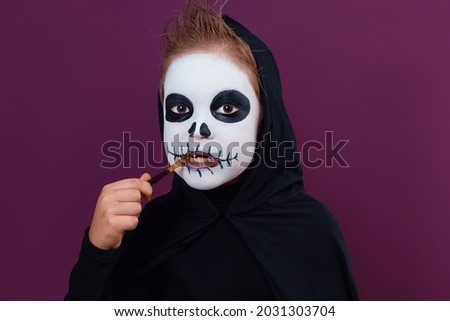 Child with Halloween makeup in skeleton costume eats Halloween jelly worms on dark purple background.