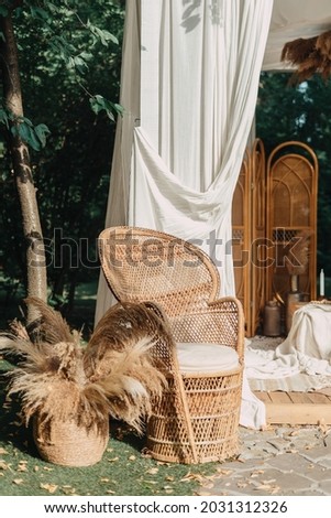 Backyard arbour design, natural decoration outdoor. Cozy rural style for trendy veranda.Wicker furniture, armchair near dry plant in brown basket at green garden background.