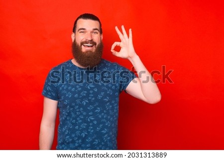 Bearded hipster wearing a blue T-shirt is showing ok gesture over red background.