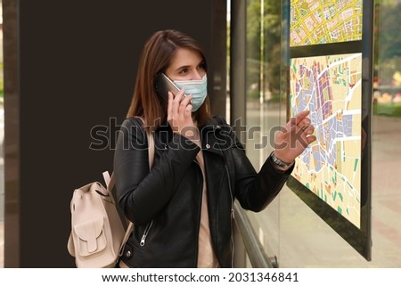Young woman in medical face mask talking on phone near public transport map at bus stop