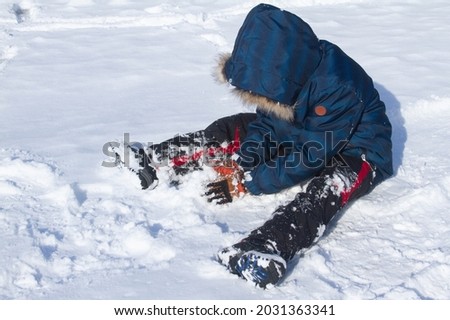 A boy in winter blue clothes sits in the snow and plays with snow, winter fun, snowfall, cold frosty day, children's winter games, morning silence in the mountains, children's tourism.
