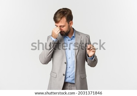 Portrait of tired male entrepreneur in grey suit, taking-off glasses and rubbing eyes, standing over white background
