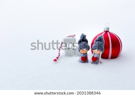 Wooden figures of boy and girl with white porcelain train and red tree bauble in the background in snow. Copy space, selective focus. Christmas card. 