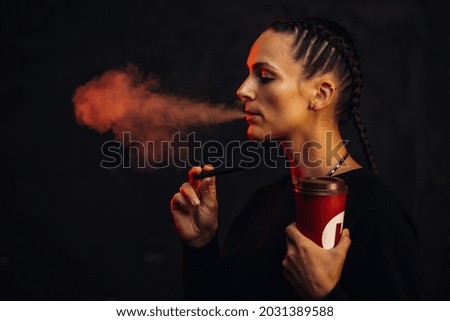 A beautiful young woman in black background with backlight smokes an electronic cigarette. Girl vaping at the bar