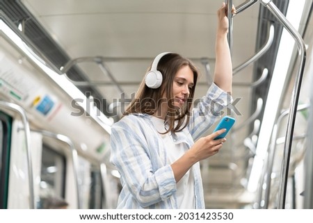 Young girl listen music in headphones and hold smartphone in subway car travel underground use wireless internet connection. Happy female chatting online in mobile phone messenger watch video in metro Royalty-Free Stock Photo #2031423530
