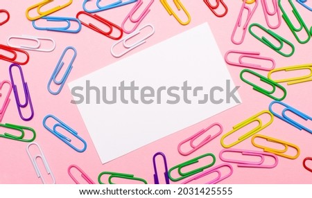 On a light pink background, colorful bright paper clips and a white blank card with a place to insert text in the center. Template. Copy space