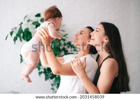 Two beautiful young women in casual clothes hug and look at their little child in the apartment. the concept of lgbt people. lesbian marriage and adoption, homosexuals-a lesbian couple. Royalty-Free Stock Photo #2031458039