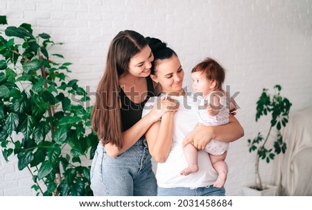 Two beautiful young women in casual clothes hug and look at their little child in the apartment. the concept of lgbt people. lesbian  marriage and adoption, homosexuals-a lesbian couple. Royalty-Free Stock Photo #2031458684