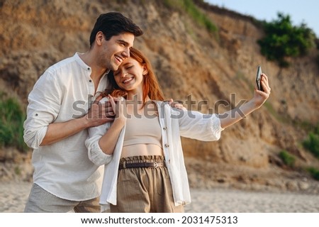 Young lovely couple two family man woman 20s in white clothes hug rest together do selfie shot mobile cell phone post photo social network at sunrise over sea sand beach outdoor seaside in summer day.