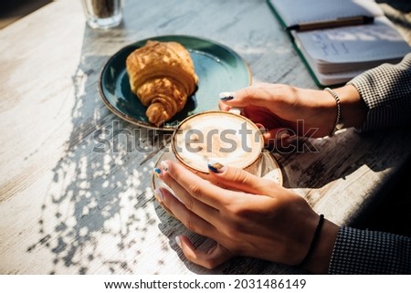 Cappuccino and croissant on the table in the cafe. The morning sunlight falls on the table, beautiful shadows appear. Delicious breakfast.Women's hands hold a cup of coffee. Royalty-Free Stock Photo #2031486149