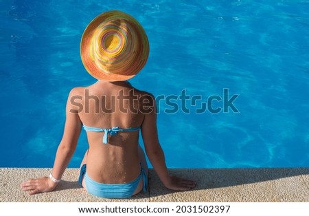 The girl sits with her back to the camera at the edge of the pool. The blue swimsuit matches the color of the pool water. Bright multi-colored hat. There is room for text. Summer positive postcard