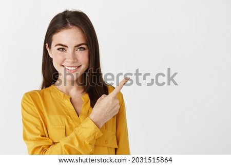 Close-up of brunette female model showing promo offer, pointing finger right and smiling at camera, white background Royalty-Free Stock Photo #2031515864