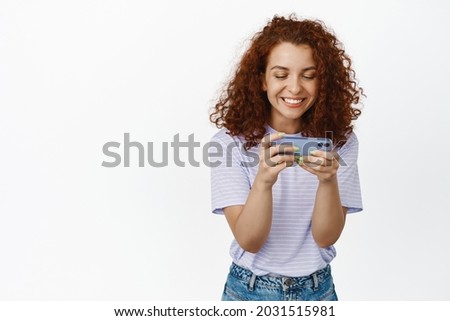 Happy redhead woman smiling, playing video game on mobile phone, holdig smartphone horizontally, watching smth on cellular internet, white background
