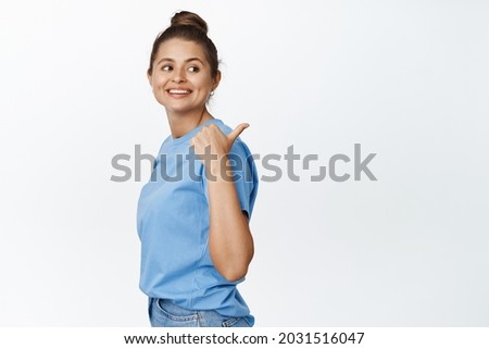 Young modern girl pointing finger right and looking behind her shoulder, showing logo advertisement, white background, wearing blue tshirt