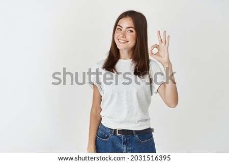 Young confident woman standing on white background and showing okay sign, confirm and approve something, praise good choice