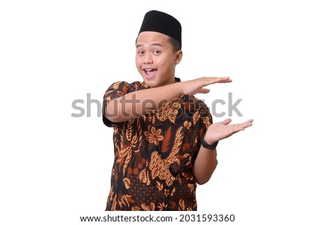 Portrait of smiling Asian man wearing batik shirt and songkok showing something with two hands. Advertising concept. Isolated image on white background