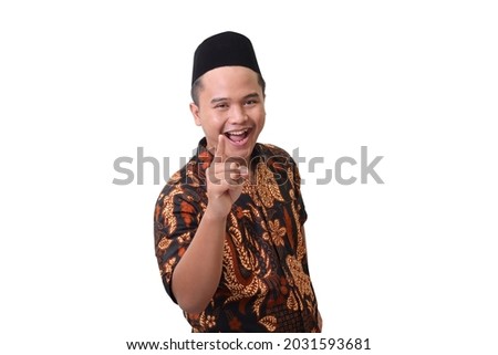 Portrait of smiling Asian man wearing batik shirt and songkok pointing his finger forward to camera. Isolated image on white background