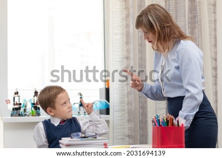 A beautiful young mother with her son is doing homework at a desk at home against a light background of a window during a pandemic quarantine. Selective focus. Portrait