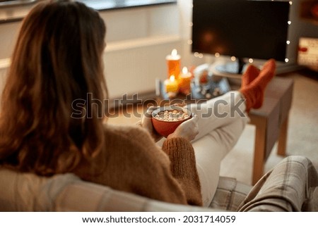 halloween, holidays and leisure concept - young woman watching tv and drinking hot chocolate with marshmallow with her feet on table at cozy home