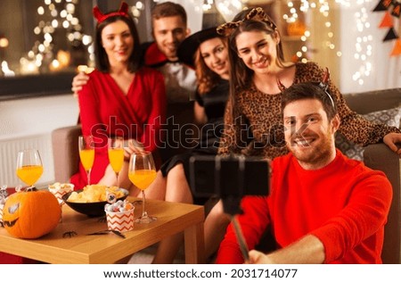 friendship, holiday and people concept - group of happy smiling friends in halloween costumes of vampire, devil, witch and cheetah taking picture by smartphone selfie stick at home party at night