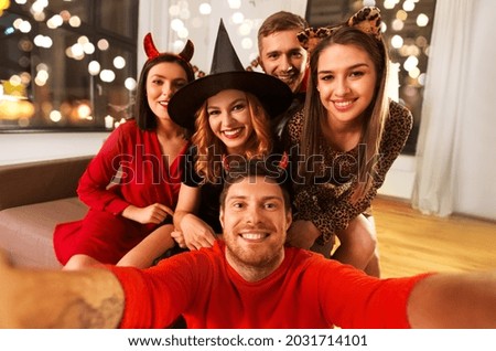 friendship, holiday and people concept - group of happy smiling friends in halloween costumes of vampire, devil, witch and cheetah taking selfie at home party at night