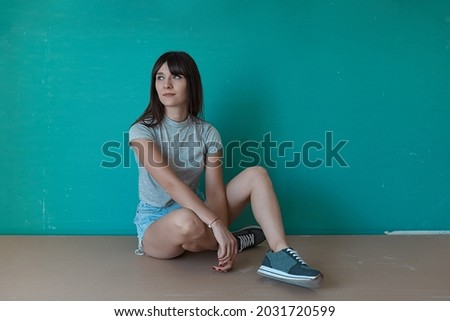 Young beautiful girl enjoy moment sitting crossed legs on a turquoise background, looking to right with copy space. Hipster girl in jeans shorts, t-shirt and sneakers.