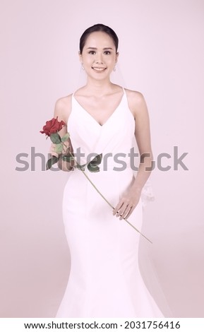 Portrait of young attractive Asian woman wearing white wedding gown holding red rose against white background. Concept for pre wedding photography.