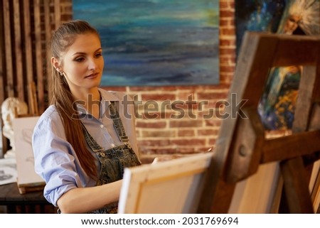 Young beautiful female artist painting on canvas using oil paintings and art brush. Painter creating artwork in art studio. Relaxation, leisure, hobby, stress management