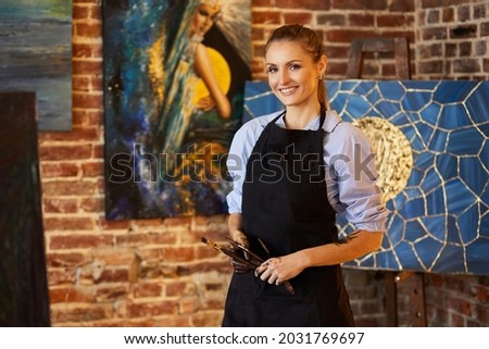 Portrait of young smiling female artist wearing apron in art studio. Painter holding art brushes, hands are stained with paint. Creative process, relaxation, leisure, hobby, stress management.