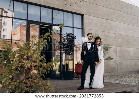 Full-length portrait of stylish newly married European couple. The bride in white dress and veil is hugging the groom. Groom with a beard and sunglasses dressed in a classic black suit. Royalty-Free Stock Photo #2031772853