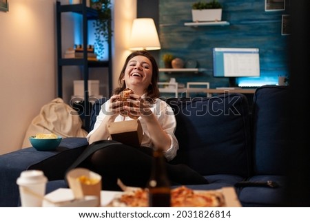 Caucasian female laughting while watching entertainment movie series on television eating tasty delicious fast food burger. Woman enjoying unhealthy takeaway food home delivered. Lunch meal order Royalty-Free Stock Photo #2031826814