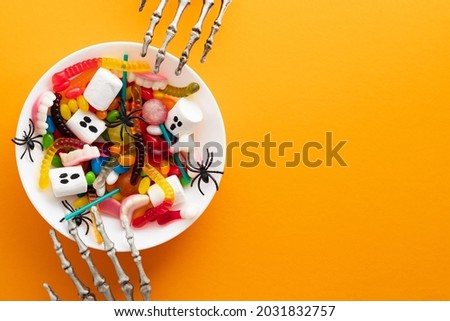 Halloween candy bowl and skeletons hands on orange background. Flat lay, top view, copy space.