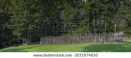 A panoramic of a garden consisting of various item such as vegetation and flowers all surrounded by a picket fence. Out to the side is an old outhouse.