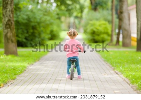 Little girl riding on first bike without pedals on sidewalk at city park in summer day. 2 years old toddler. Back view. Learning to keep balance. Royalty-Free Stock Photo #2031879152