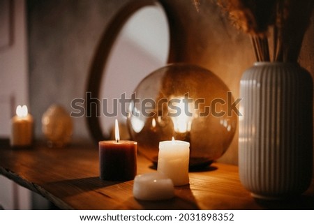 Round mirror on wooden shelf with two burning candles and small porcelain Buddha head reflected in corner of mirrored surface over concrete wall background in minimalist modern apartment interior