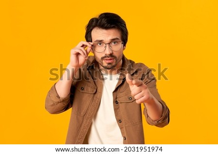 Young bearded man in glasses shows a finger, saying "you", pointing at the camera is isolated on an orange background.