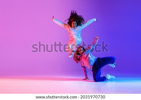Two young beautiful girls making hip-hop tricks dancing on gradient pink purple neon background. Sport achievement, expression. Concept of dance, youth, hobby, dynamics, movement, action, ad Royalty-Free Stock Photo #2031970730