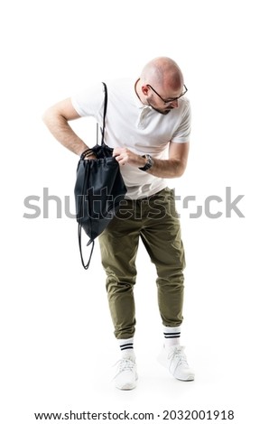 Puzzled young bald shaved nerdy man searching bag looking down for forgotten or lost item. Full length portrait isolated on white background