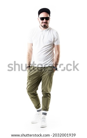 Happy comfortable stylish young man wearing chino pants cap and sunglasses smiling. Full length portrait isolated on white background Royalty-Free Stock Photo #2032001939