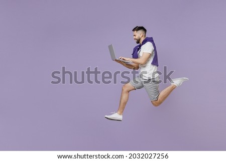 Full size body length side view profile smiling fun young brunet man 20s wears white t-shirt purple shirt jump hold use work on laptop pc computer isolated on pastel violet background studio portrait.