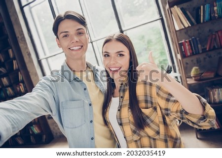Self-portrait of two attractive cheerful people hugging showing thumbup advert at library loft industrial interior indoors